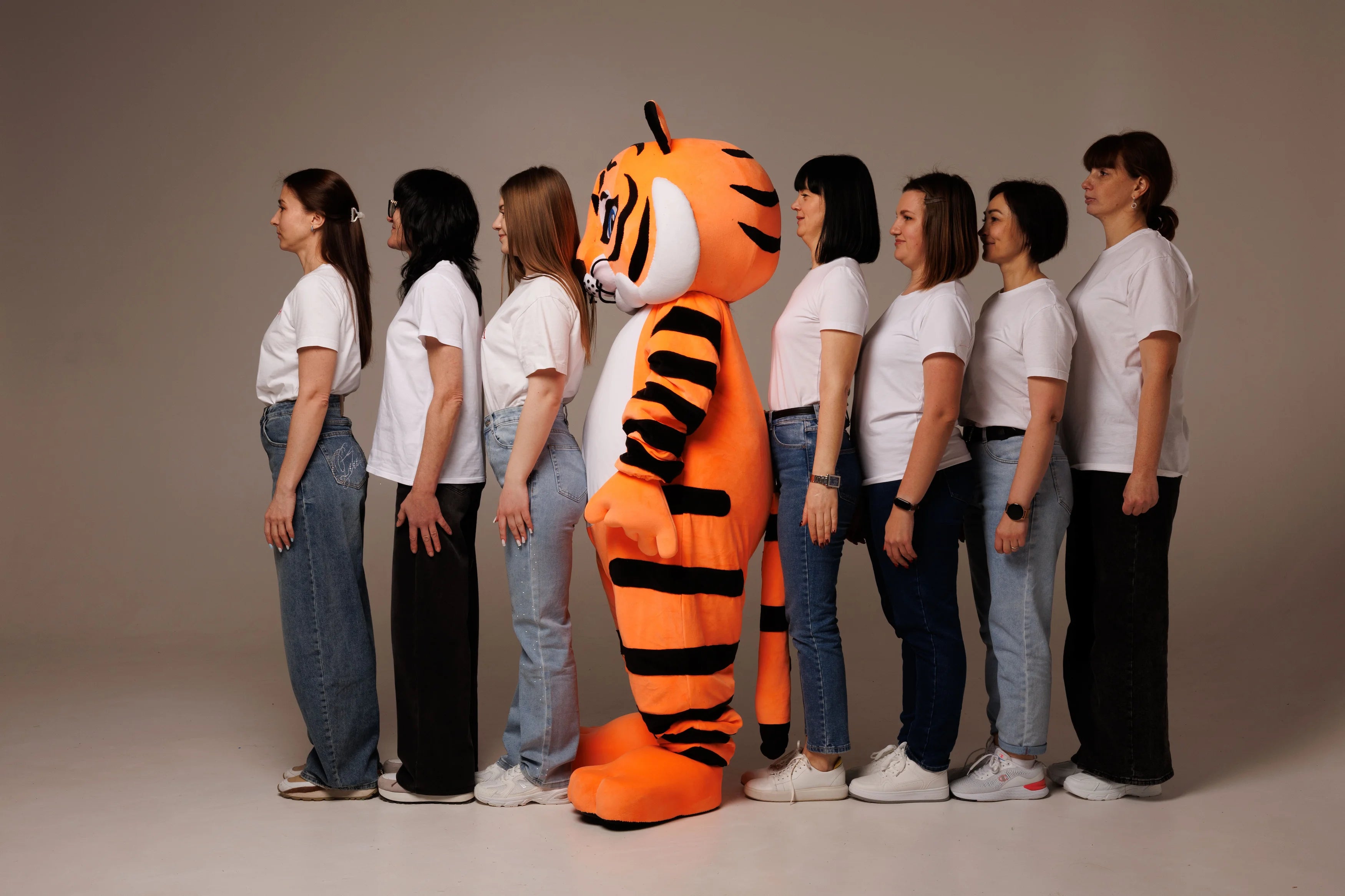 Person in a tiger mascot costume standing among people wearing white shirts and jeans on a plain background