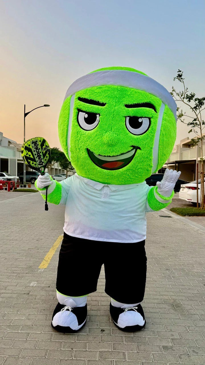 Person in a green and white mascot costume standing on a sidewalk with buildings and cars in the background.